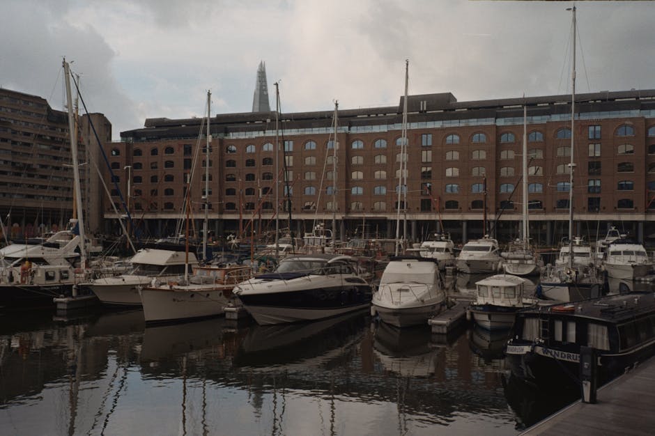 A marina with multiple yachts and boats docked along wooden piers in front of a large brick building with arched windows, located in an urban waterfront area. The boats vary in size and design, with s
