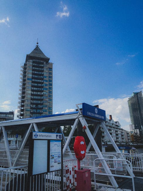 A marina with multiple yachts and boats docked along wooden piers in front of a large brick building with arched windows, located in an urban waterfront area. The boats vary in size and design, with s