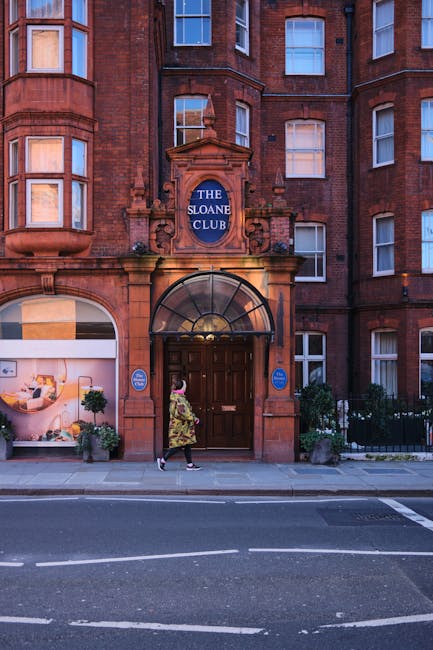 A woman walking past the entrance of The Sloane Club, a red-brick residential building with white-framed windows and a central arched doorway. The building features a blue sign with white letters indicating the club's name above the entrance, which is framed by decorative stonework and a glass canopy. The sidewalk in front has potted plants on either side of the doorway, and a woman wearing a floral dress and carrying a bag is approaching or leaving the building. The scene is set on a street in Chelsea, with a pedestrian crossing visible in the foreground and the building illuminated by natural daylight. This setting is relevant to house removals and furniture transport services offered by Chelsea Removals, often involving packing, loading, and relocating household items in the area.