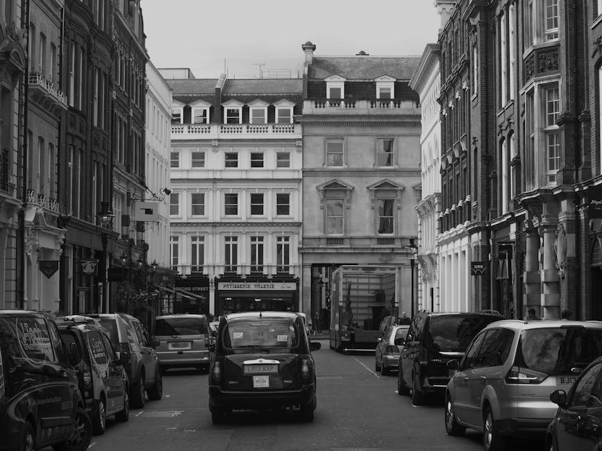 A narrow street in Chelsea lined with multi-storey Victorian and Georgian-style buildings featuring ornate facades, tall sash windows, and decorative architectural details. Several parked cars, including a small black vehicle at the center and other larger vehicles on the pavement, are visible along both sides of the street. In the background, trucks and vans are parked near the building entrances, with one van positioned at the curb, likely involved in a home relocation or furniture transport process. The street appears to be in a commercial or residential area, with a parking area filled with vehicles and a storefront with visible signage. The scene is captured in black and white, emphasizing textures, architectural details, and the spatial arrangement typical of central London neighborhoods. Chelsea Removals could be engaged in loading or unloading furniture and boxes as part of their movers' activities, supporting efficient packing and moving logistics for clients in the area.